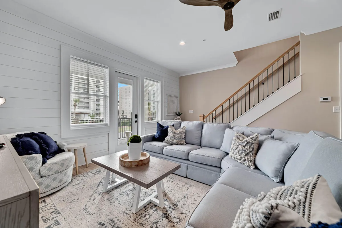 A cozy living room featuring a large sectional sofa and a wooden coffee table, set near a staircase. The inviting atmosphere is designed for relaxing, socializing, or reading, and the space is filled with warm, natural light that hints at a tranquil retreat ideal for extended stays.