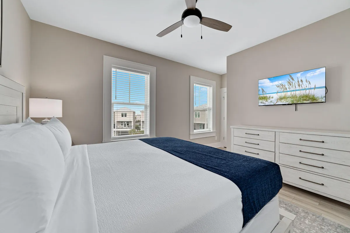A serene bedroom featuring a bed with crisp white and navy bedding, two windows with blinds letting in natural light, a wall-mounted television, and a dresser with multiple drawers. The space feels inviting and modern, perfectly suited for relaxing and unwinding at the end of the day.