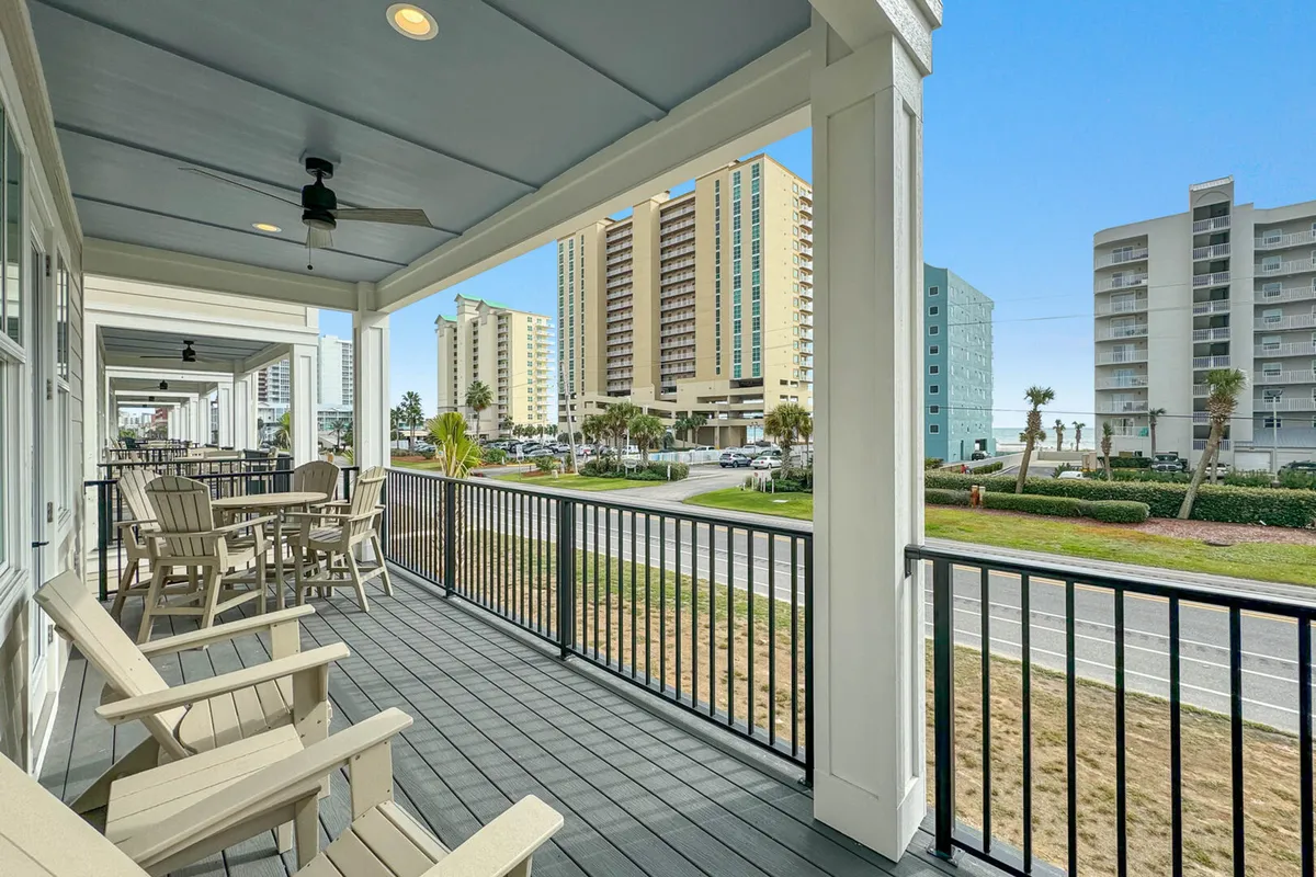 The image showcases a spacious, inviting balcony with comfortable seating, framed by modern railings and columns. The balcony overlooks lush greenery, a paved walkway, and offers glimpses of nearby high-rise buildings, hinting at proximity to both vibrant community life and tranquil beach surroundings.