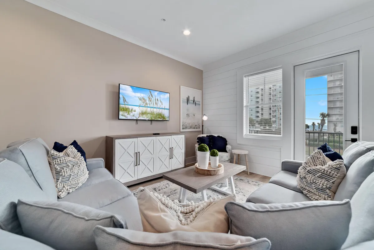 A sun-filled living room with soft gray sofas and textured pillows faces a wall-mounted flat screen TV displaying a beach scene. A wooden coffee table with decorative plants adds warmth, while the glass door and window offer views of palm trees and nearby buildings, hinting at the private balcony access.