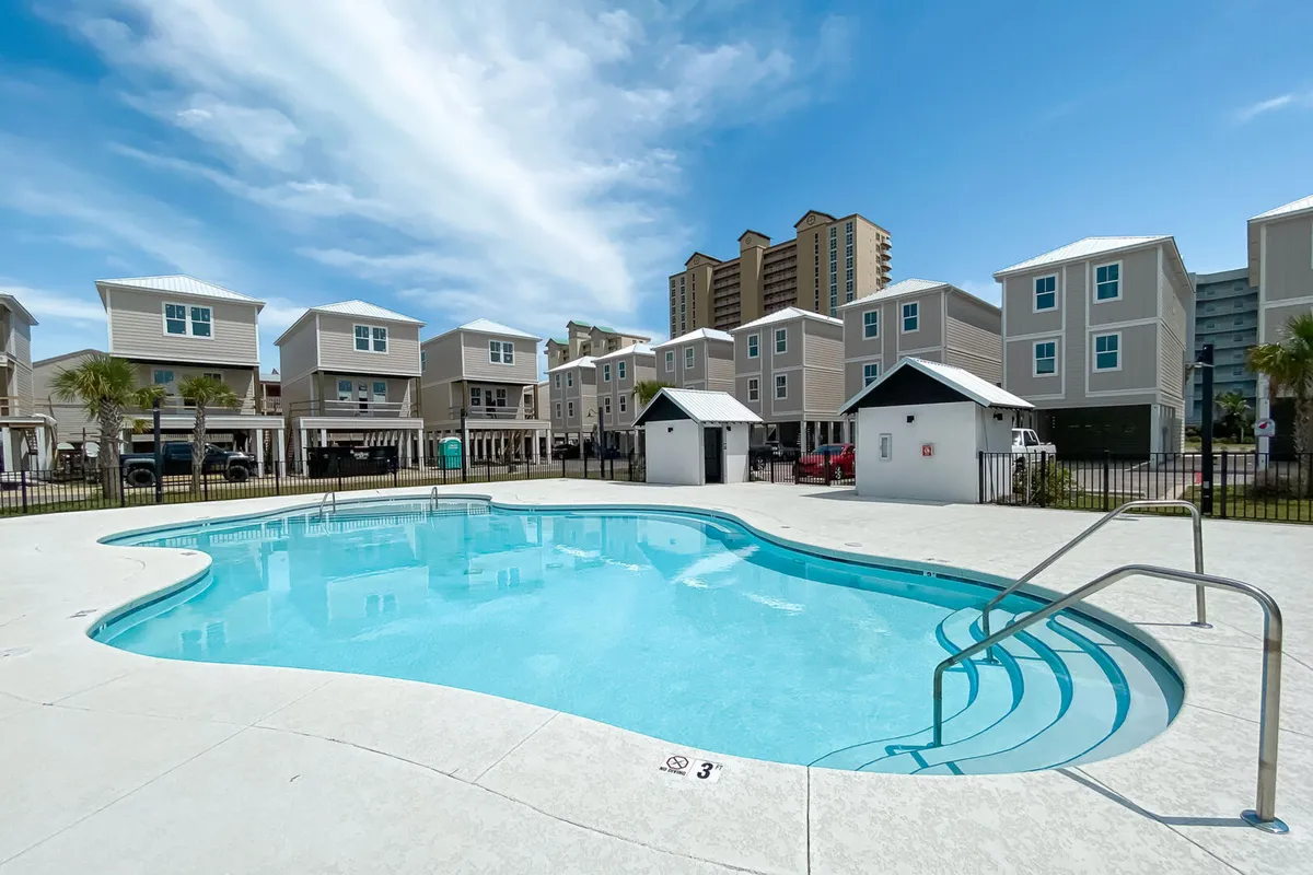 A sparkling community swimming pool is set against a backdrop of modern stilted beach homes. The pool features curved edges, steps for easy entry, handrails, and depth markers. The blue sky and light clouds overhead evoke classic vacation vibes, while the scene suggests both relaxation and family fun just steps from the beach.