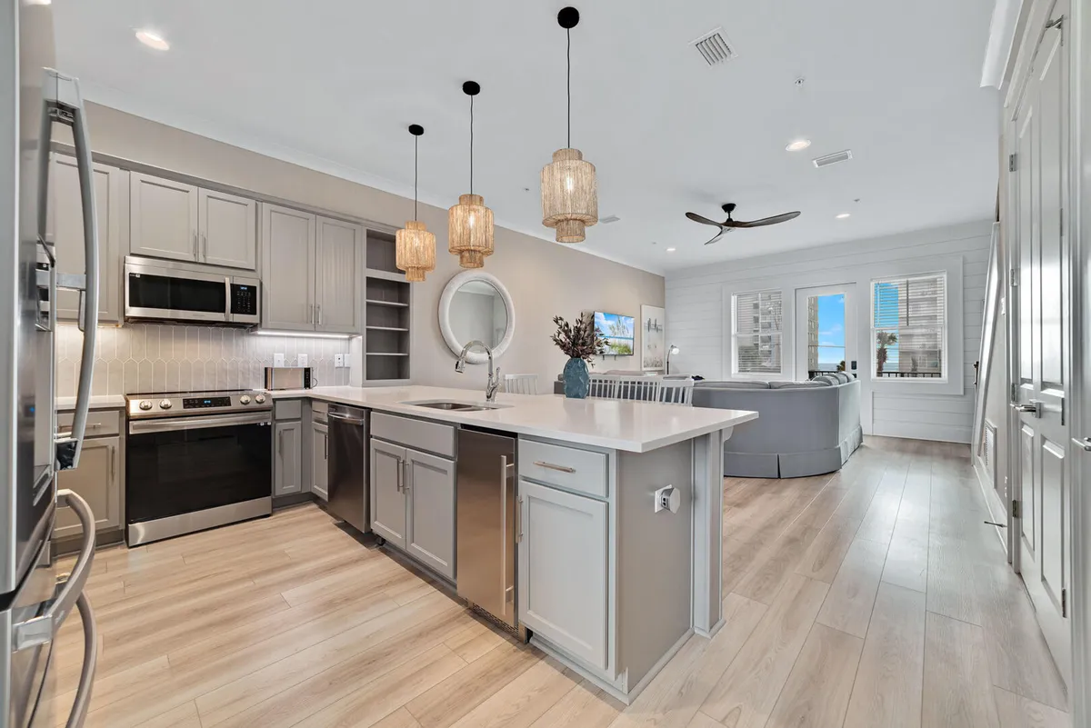 The image presents a contemporary open-plan space with a kitchen island and pendant lighting, stainless steel appliances, a large L-shaped sofa, and a visible smart TV in the living area. The setting invites communal cooking, dining, and relaxing, with a clear flow between kitchen, dining, and living spaces.