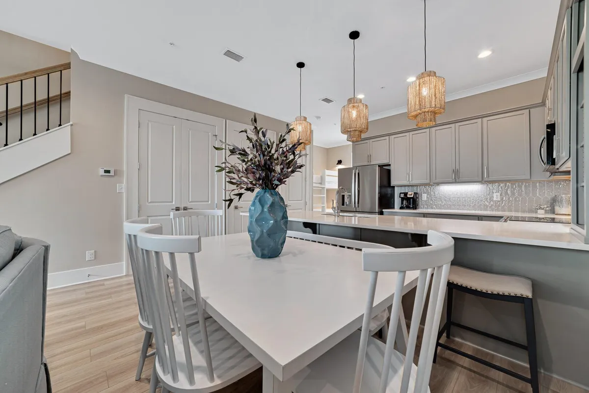 This image features an inviting, modern kitchen and dining area with a white dining table adorned with a blue vase and floral arrangement. The kitchen boasts a spacious island with elegant pendant lights, stainless steel appliances, and a staircase leading to the upper floor. Natural light and tasteful decor create a calm, welcoming atmosphere.