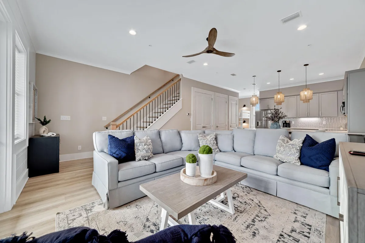 This image features a large, light-gray sectional sofa with decorative pillows at the center of a spacious, open-plan living area. The wooden coffee table with small plants creates a cozy, inviting centerpiece. In the background, a modern kitchen with pendant lighting and bar seating flows seamlessly into a dining area with seating for six, all bathed in natural light.