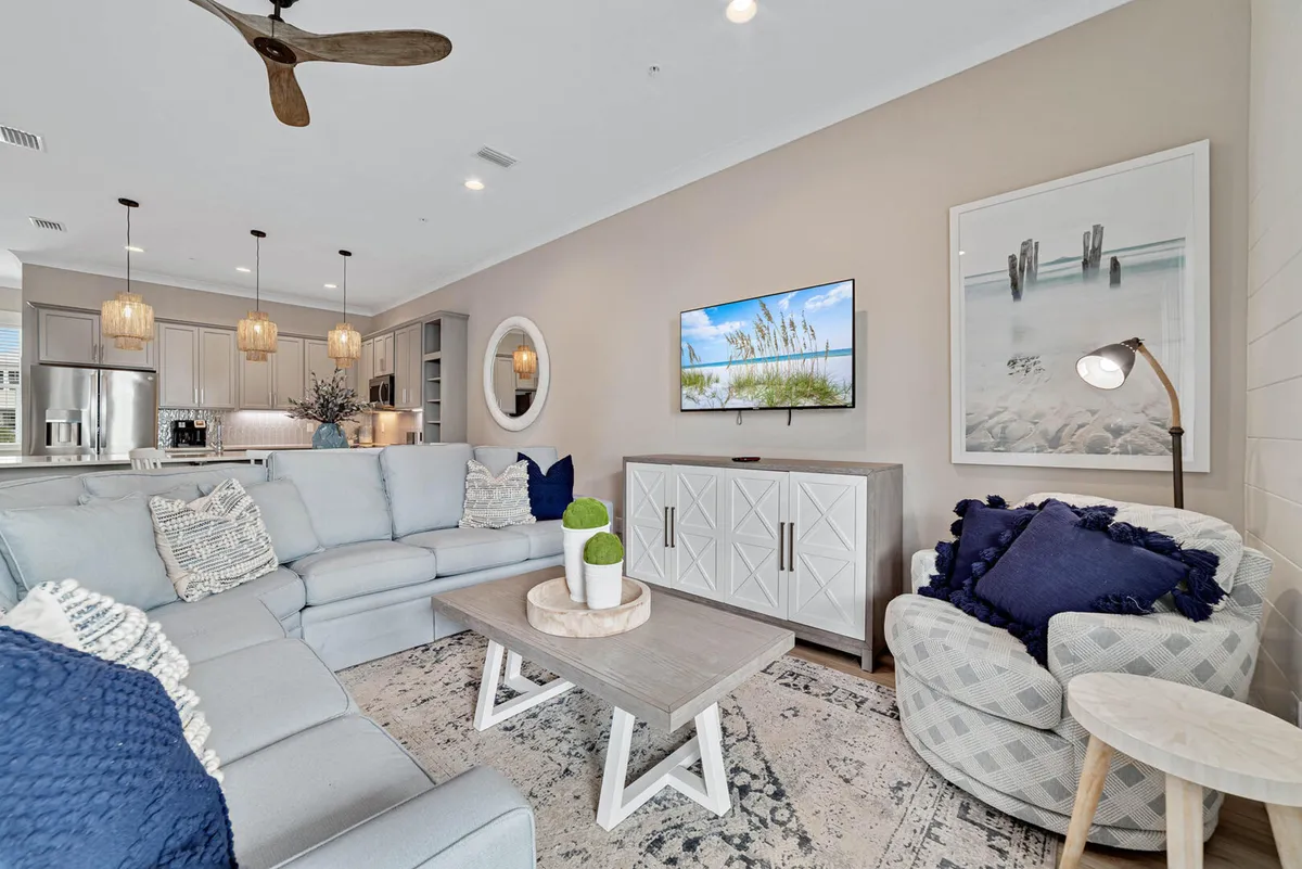 This image features a spacious living area with a large sectional sofa and an open kitchen, all bathed in natural light. The glass doors and windows suggest a strong visual connection to the outdoors, including the beach or lagoon area nearby. The mounted TV displays a beach scene, reinforcing the home's connection to water activities and the surrounding coastal environment.