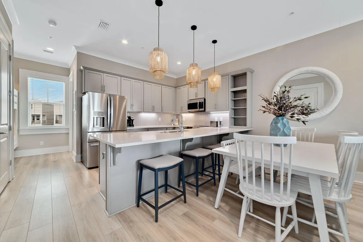 Bright and airy kitchen with a large island and seating, stainless steel refrigerator, white dining table with chairs, and stylish decor including a round mirror and vase. The atmosphere is lively and social, inviting casual dining and connection.