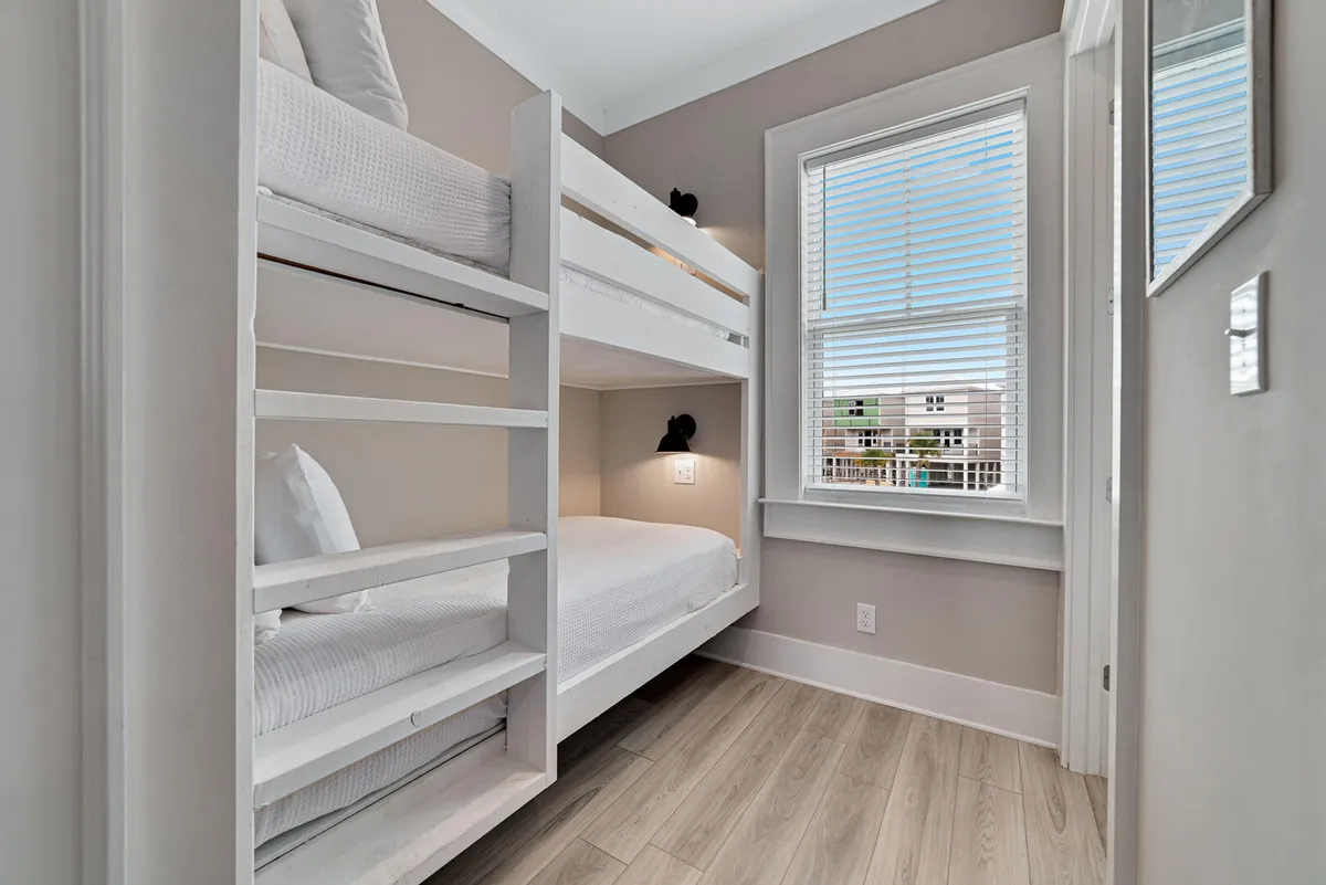 A cozy bunk area featuring white wooden twin bunk beds, a large window with white blinds letting in natural light, and a wall-mounted reading light for each bed. The arrangement creates an inviting and peaceful nook perfect for children or extra guests.