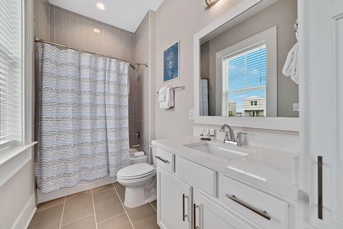 A bright and spotless bathroom featuring a white vanity with sink and chrome faucet, a clean toilet next to a bathtub with a striped shower curtain, and a window letting in ample natural light. The space radiates freshness and order, evoking a hygienic and welcoming atmosphere.