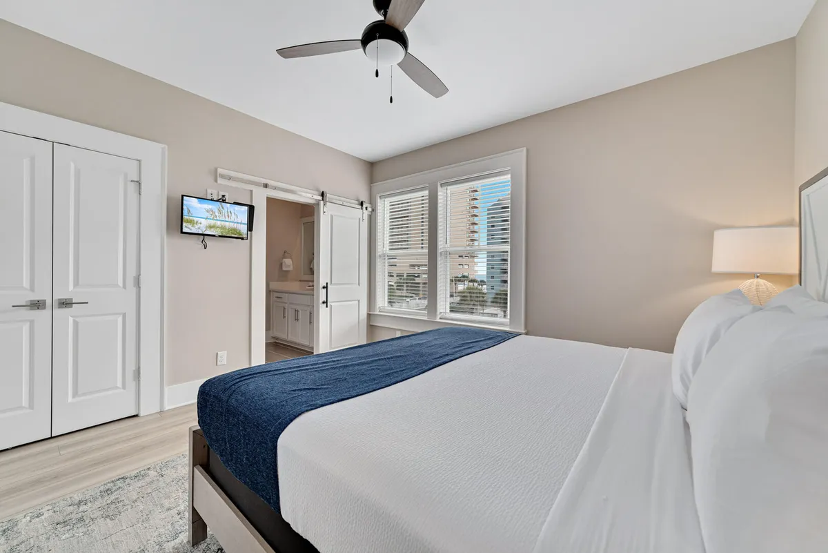 A peaceful guest bedroom with a king size bed dressed in white linens and a navy throw, a window allowing in natural light with views of neighboring buildings, a barn door leading to a private bathroom, and a wall-mounted TV. The layout emphasizes comfort and privacy.