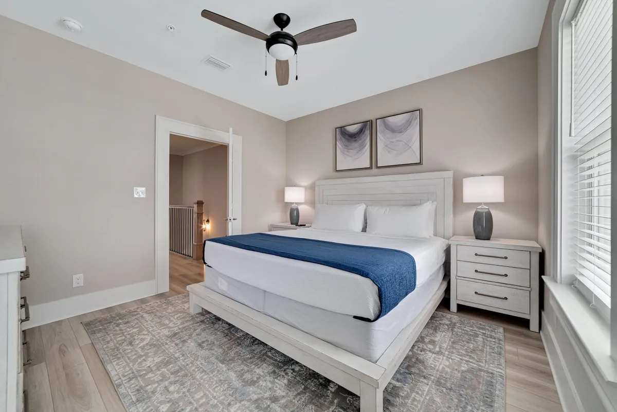 A bright, airy bedroom featuring a king-size bed with a blue throw, bedside tables with lamps, and abstract art above the bed. The space exudes tranquility and is exceptionally tidy, with natural light streaming in.