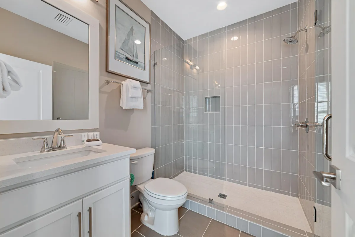A modern bathroom with a glass-enclosed walk-in shower, white vanity with integrated sink and mirror, and nautical-themed artwork above a towel rack. The space feels calm, orderly, and meticulously maintained.