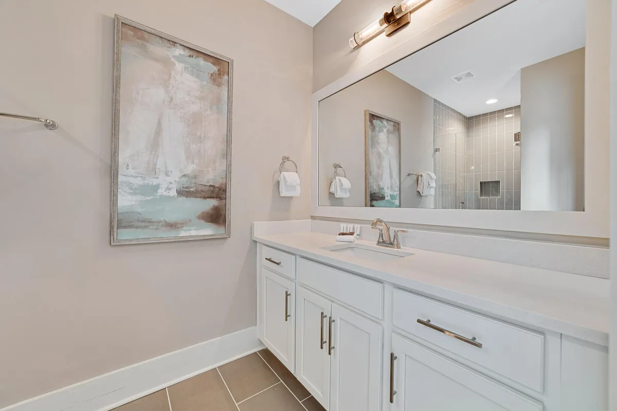 A serene bathroom with a large mirror above a white vanity, multiple storage cabinets, and a glass-enclosed shower reflected in the mirror. The overall aesthetic is bright, fresh, and meticulously maintained.
