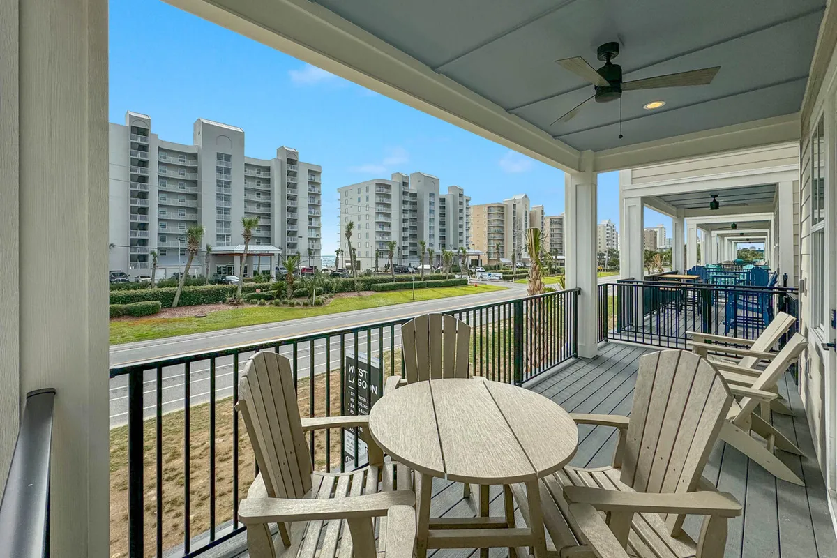 A covered balcony with wooden chairs and a round table overlooks a row of similar balconies and distant multi-story buildings. The space is shaded and inviting, perfect for morning coffee, casual dining, or sunset views, and visually connects interior comfort with Gulf Coast scenery.