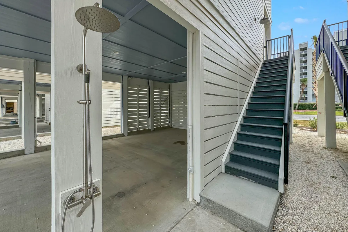 This exterior shot features a modern outdoor shower with a rain showerhead, a staircase leading to the upper level, and an open, shaded area beneath the house. The white siding and relaxed setup evoke a classic coastal vibe. The image captures the experience of rinsing off sand after a beach trip and the convenience of transitioning from outdoor adventures to private home comforts.