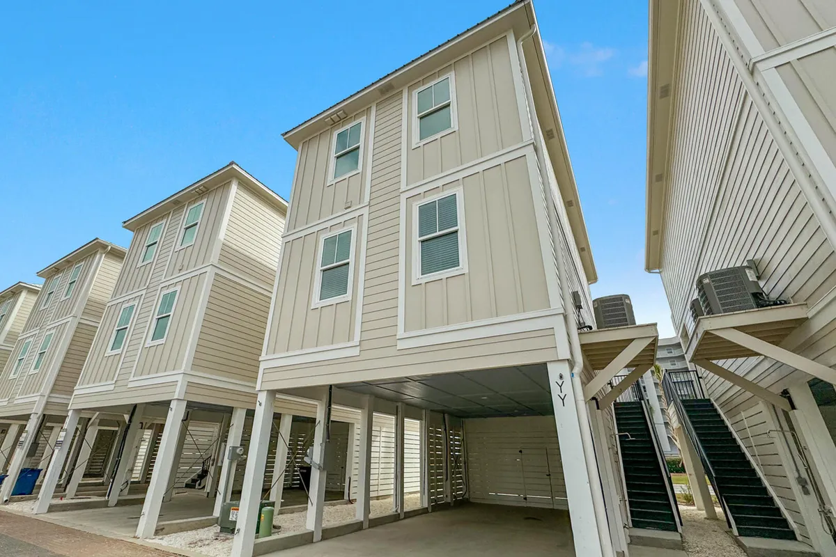 A wide view of a three-story beach house on stilts shows the covered parking area beneath, exterior stairs, and the architectural unity of neighboring homes. The scene is bright and welcoming, emphasizing the secure and convenient parking for multiple vehicles and the sense of community.