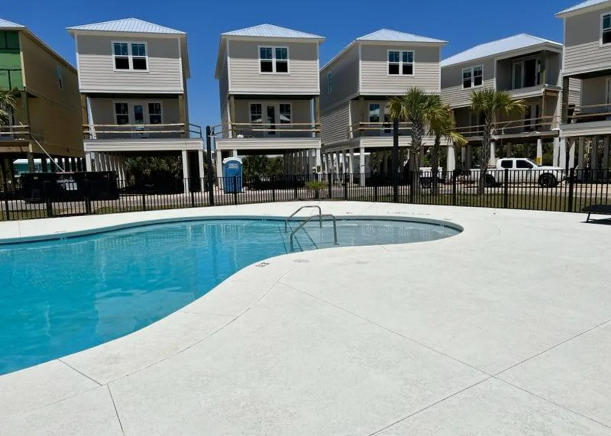 This exterior image features a sparkling community swimming pool surrounded by modern stilt homes, palm trees, and a secure fence. The clear blue water and open sky evoke relaxation and fun, setting the scene for sun-drenched swims and social gatherings within the resort community.