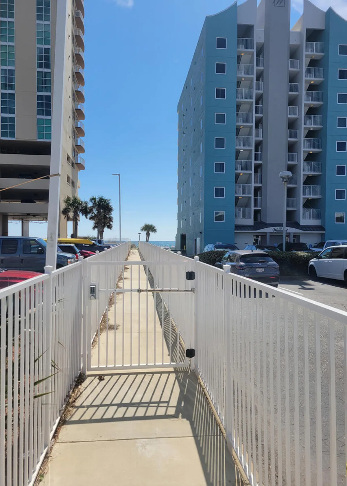 A white gated pathway leads directly to the beach, with palm trees, high-rise buildings flanking the walk, and the ocean visible on the horizon. The exclusive access gate is prominent, inviting anticipation for the beach beyond.